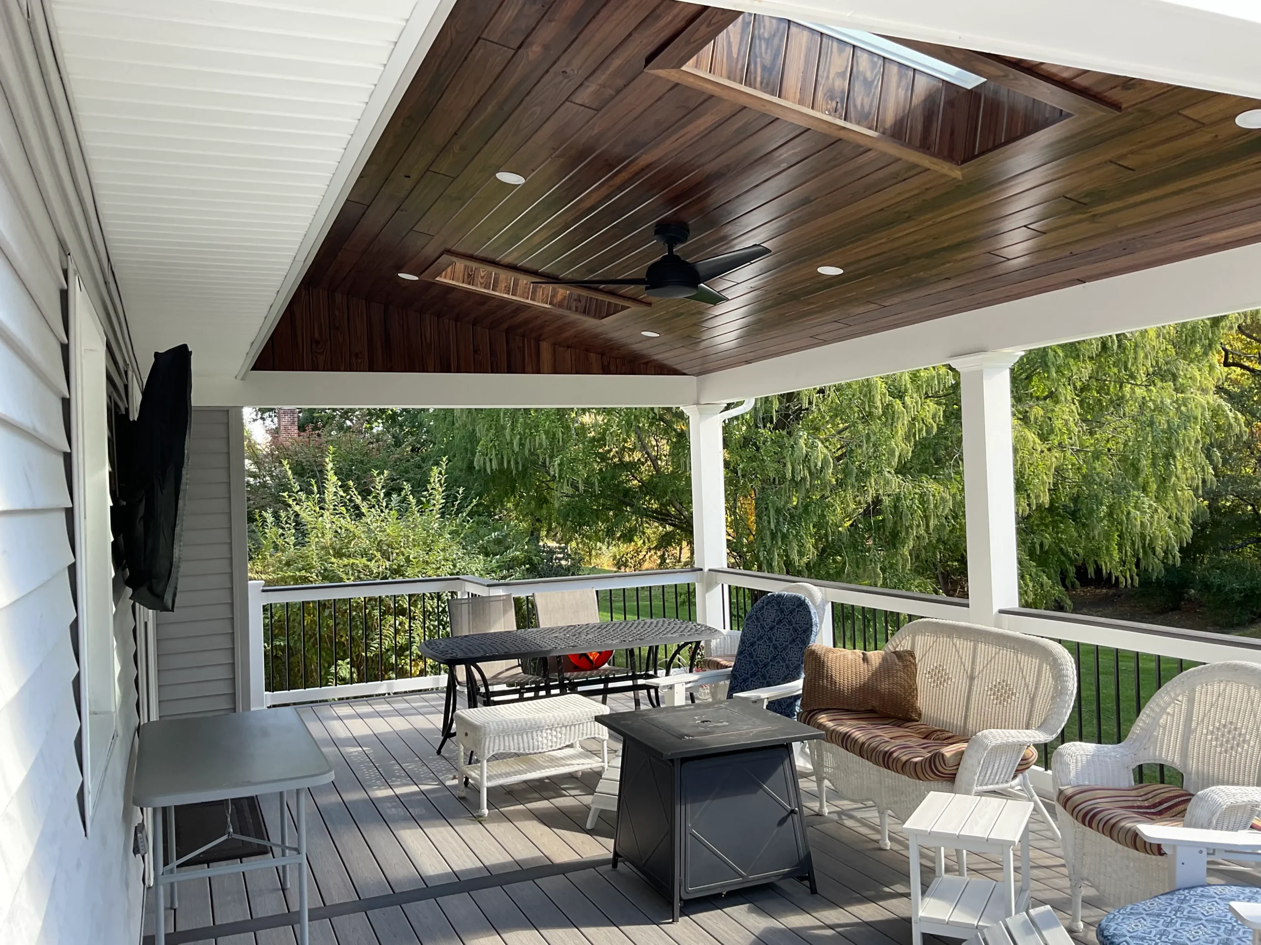 Covered back porch with dark wood ceiling, white railings, and outdoor furniture including wicker chairs and a metal table overlooking trees.