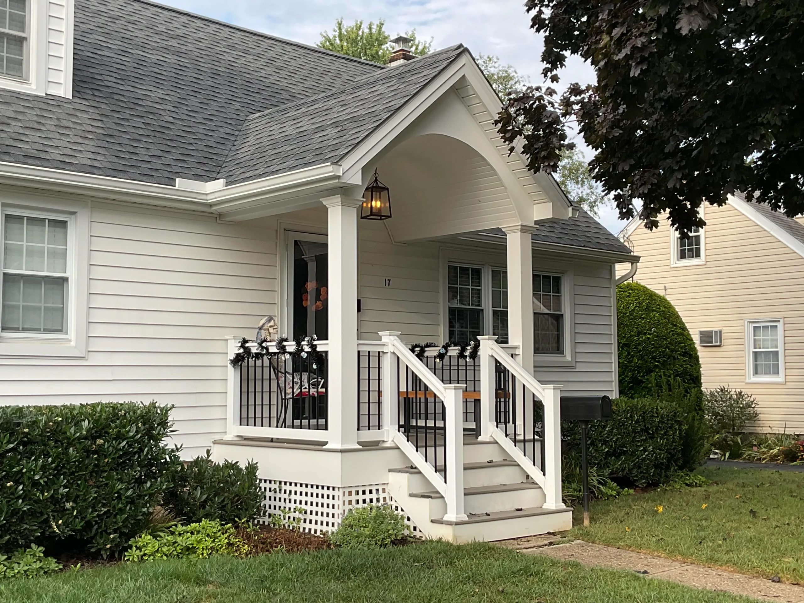 pa-exterior-updates-macungie - Skyline Exteriors - Skyline Exteriors Front porch of a light-gray house with white trim, black railing, and wooden stairs surrounded by greenery.
