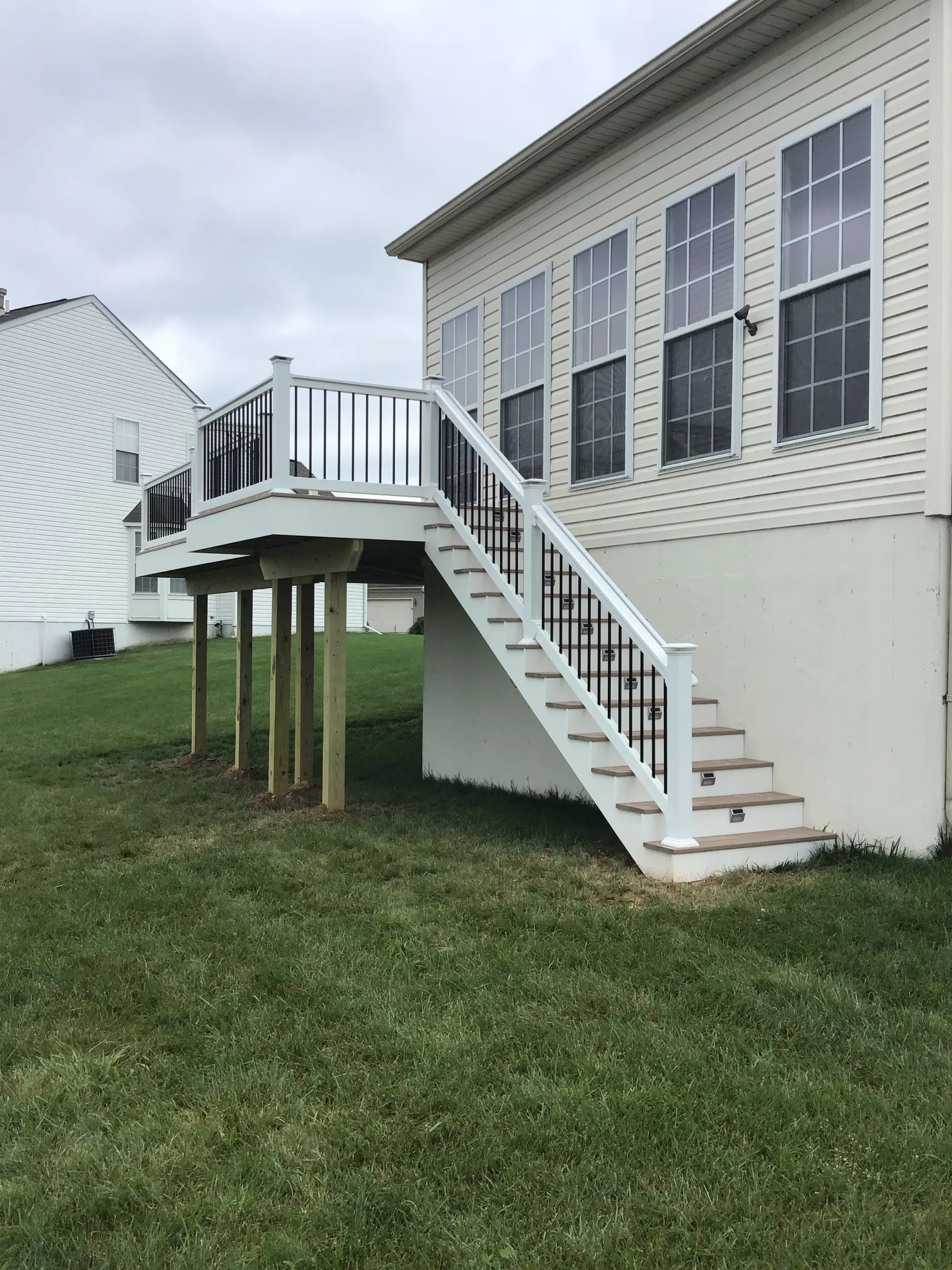 Backyard view of a beige house with an elevated white deck and stairway leading to the second floor, over a green lawn under a cloudy sky.