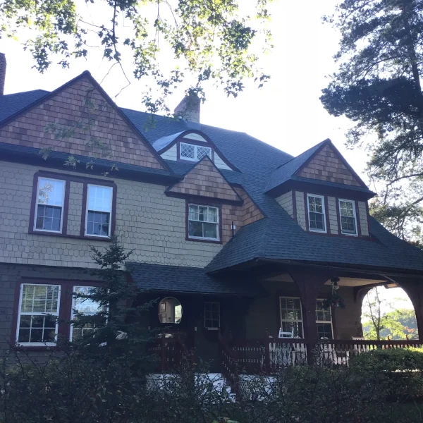 Two-story house with brown shingle siding, steep gabled roof, and brick trim, set among trees and a wooden fence in foreground, daylight.