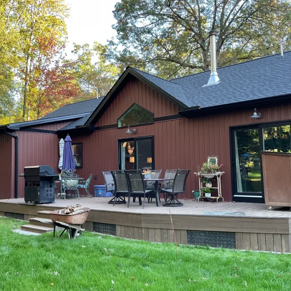 Backyard deck attached to a brown house with glass doors, outdoor dining set, grill, and potted plants; autumn trees behind.