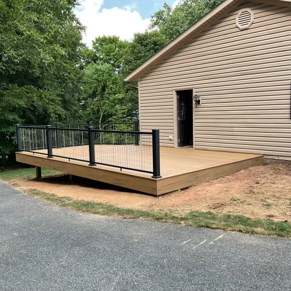 Wooden deck attached to a beige house, with black metal railing, an open door, and a window with shutters near trees and greenery.