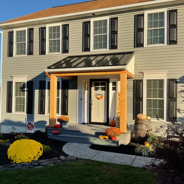 Two-story beige house with black shutters and a glass-roof porch, decorated with pumpkins and mums for fall, in a landscaped yard.