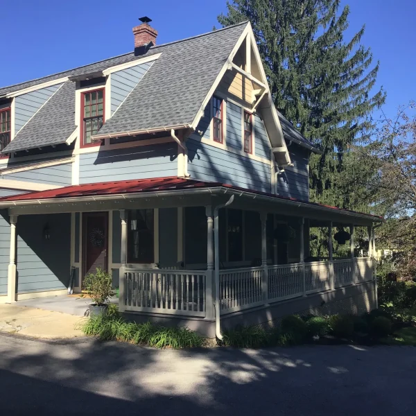 Blue two-story house with a steeply pitched roof, red-trimmed windows, and a wraparound porch on a sunny day.