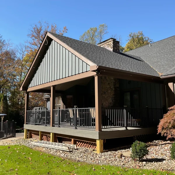 Front view of a house with a dark gray roof, covered porch, and brown trim amid fall trees under a blue sky.
