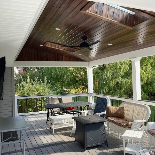 Covered outdoor deck with a dark wood ceiling and ceiling fan, white railing, and wicker seating facing greenery