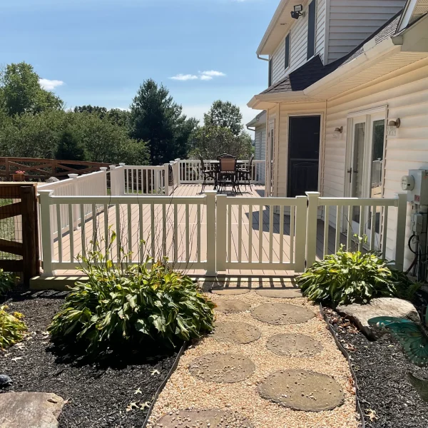 Backyard deck scene: beige house, white railing, round table with chairs, and a stone stepping-path leading to a garden area.