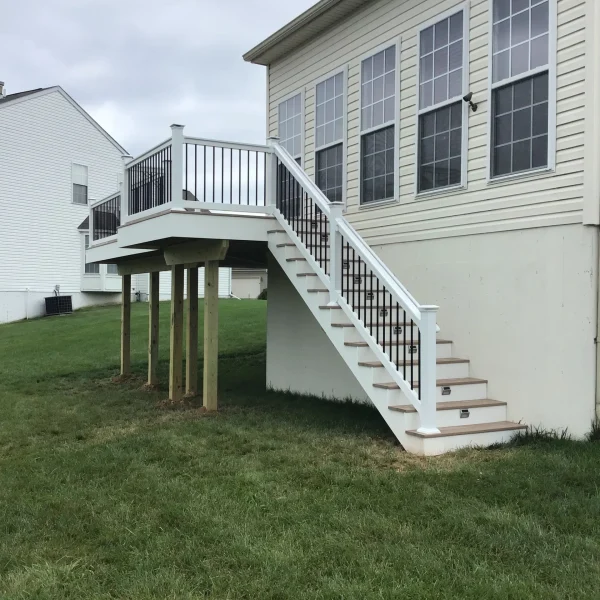 Backyard view of a beige house with an elevated white deck and stairway leading to the second floor, over a green lawn under a cloudy sky.