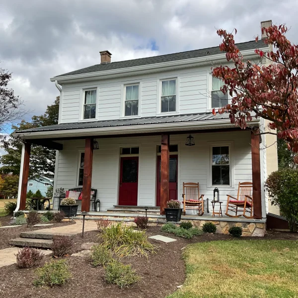 White two-story house with red front doors, a covered porch, and wooden rocking chairs; fall foliage in the yard.