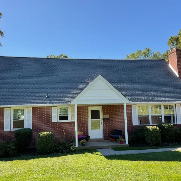 Brick suburban house with a steep dark shingled roof, white-framed windows, and a small front porch facing a green lawn under a clear blue sky.
