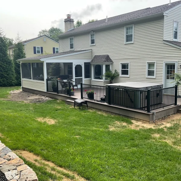 Backyard view of a beige two-story house with a screened porch and black railing deck, green lawn, and stone edging on the left.