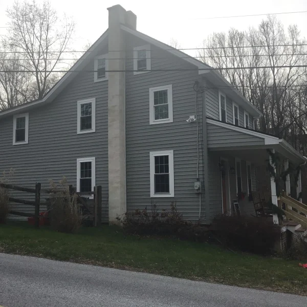 Gray two-story house with white window trim, tall chimney, and leafless trees in the background; power lines cross the scene.