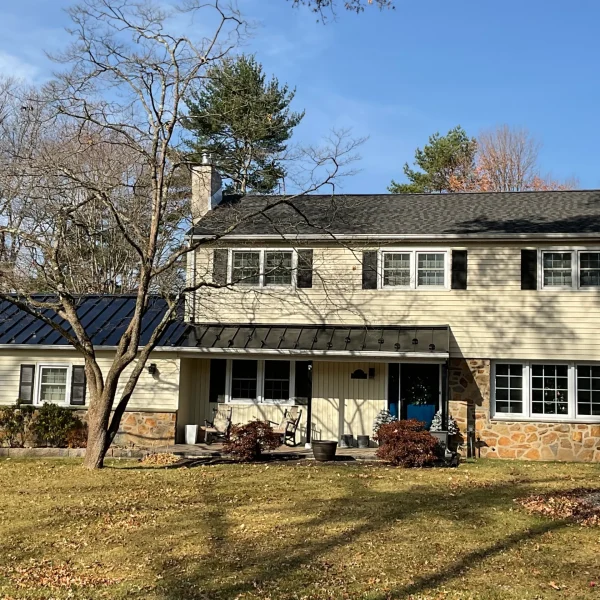 Two-story beige house with stone accents and black shutters, leafless trees in the front yard on a sunny day.