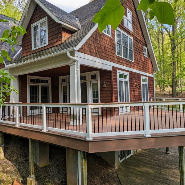 Two-story red wooden house with white trim and multiple dormer windows, surrounded by green trees and a large wooden deck.