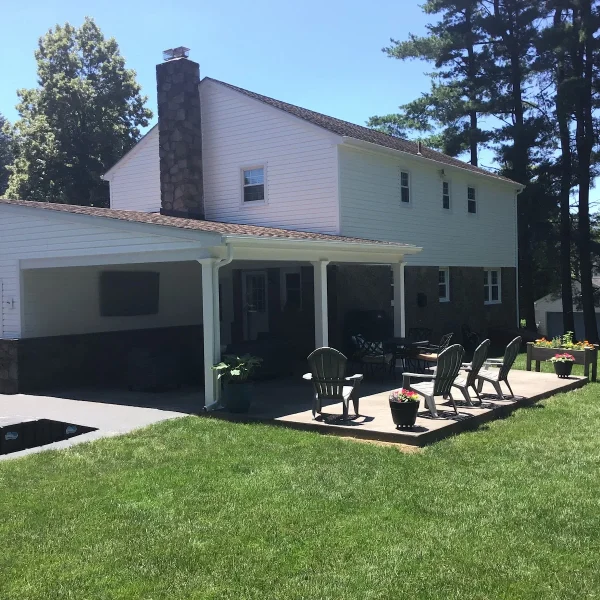 White two-story house with a stone chimney and covered patio, outdoor seating on a wooden deck, green lawn and tall trees in the background